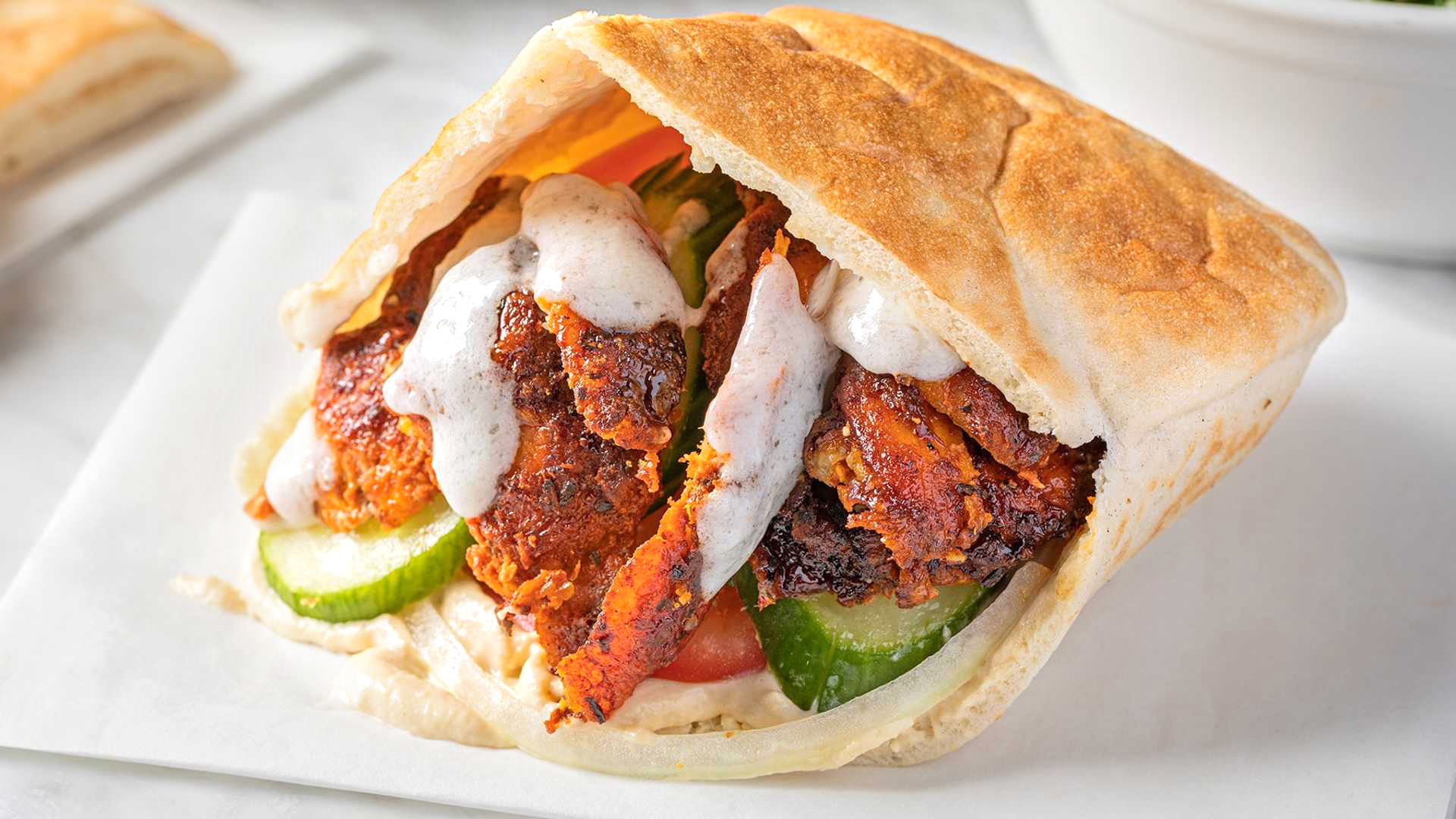 A close up of a Chicken Shawarma Sandwich from Sultan's Market. Which Consist of a warm pita lined with hummus, stuffed with marinated spit roasted chicken with onions, cucumbers, tomatoes and garlic sauce.