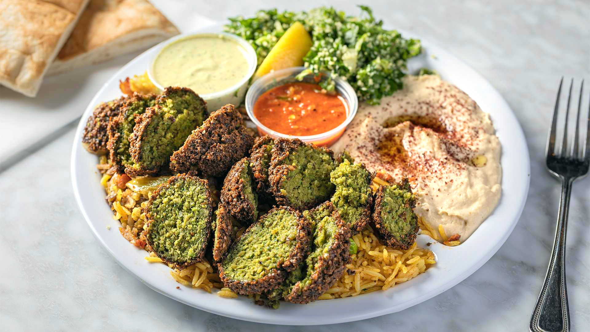 A close-up of a Falafel Dinner from Sultan's Market: Which consist in 3 pieces of falafel cut on top of your choice of (curry rice or lentils) sides of hummus, tabuli, tahini sauce, hot sauce and a pita.