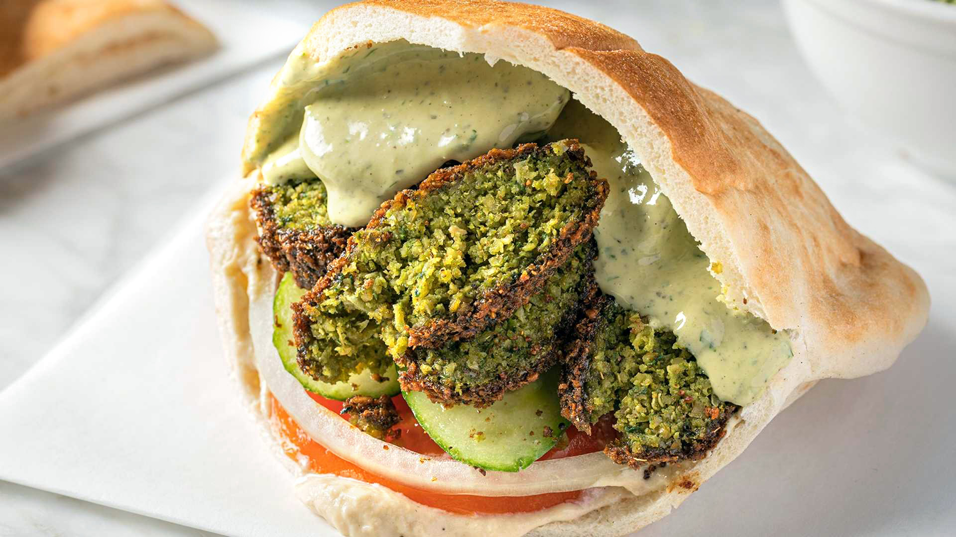A close-up of a Falafel Sandwich from Sultan's Market: Which consists of a warm pita lined with hummus, stuffed with falafel, cucumbers, tomato, onion and tahini sauce. This is a Vegan dish.