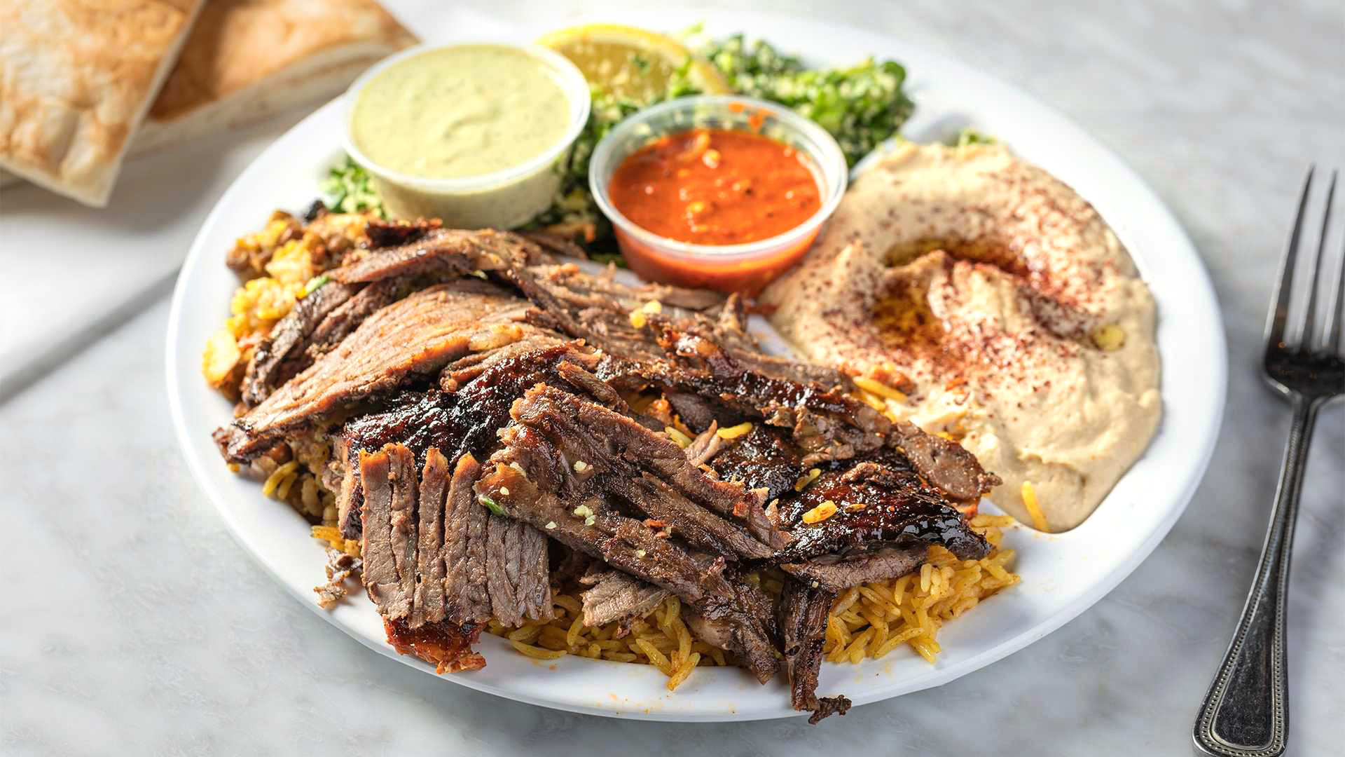 A close up of a Lamb Shawarma Dinner from Sultan's Market. Which Consist of Marinated spit roasted lamb with a side of tahini sauce, a side of rice, hummus and tabuli salad.