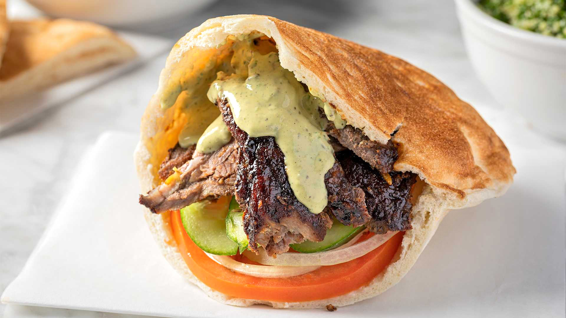 A close up of a Lamb Shawarma Sandwich from Sultan's Market. Which consist of Marinated spit roasted lamb and beef thinly sliced lamb, served in a pita with hummus, onions, cucumbers, tomatoes and tahini sauce.